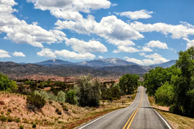 A view down the highway on the High Road, partly cloudy skies and green trees covering the high desert
