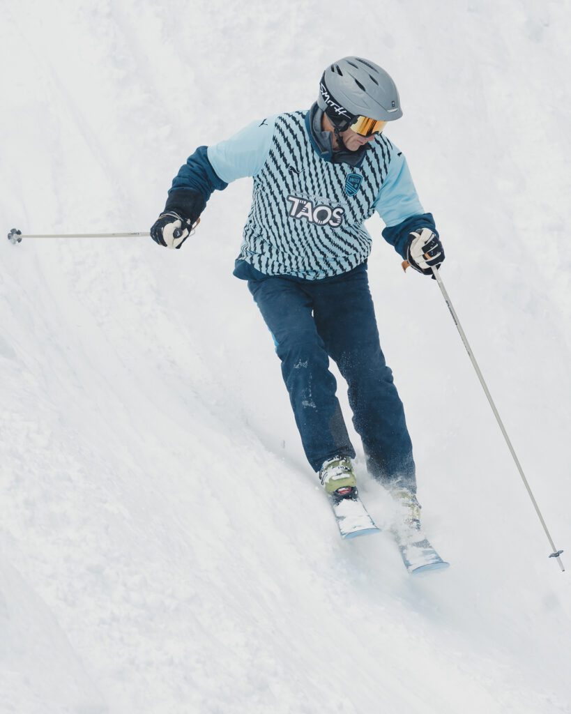 A skier wearing a striped turquoise jersey that says Visit Taos