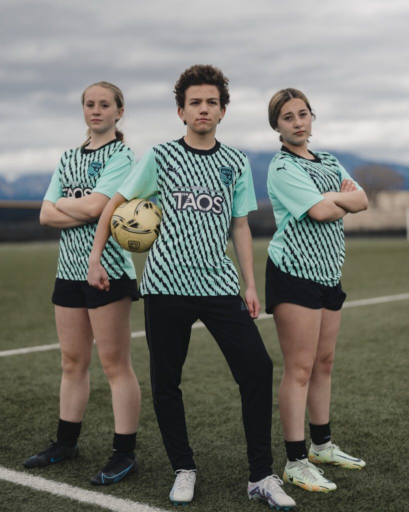Three young soccer players wearing a striped turquoise jersey that says Visit Taos