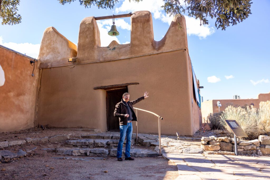 A man addresses an off-camera group of people as he stands in front of an old adobe building